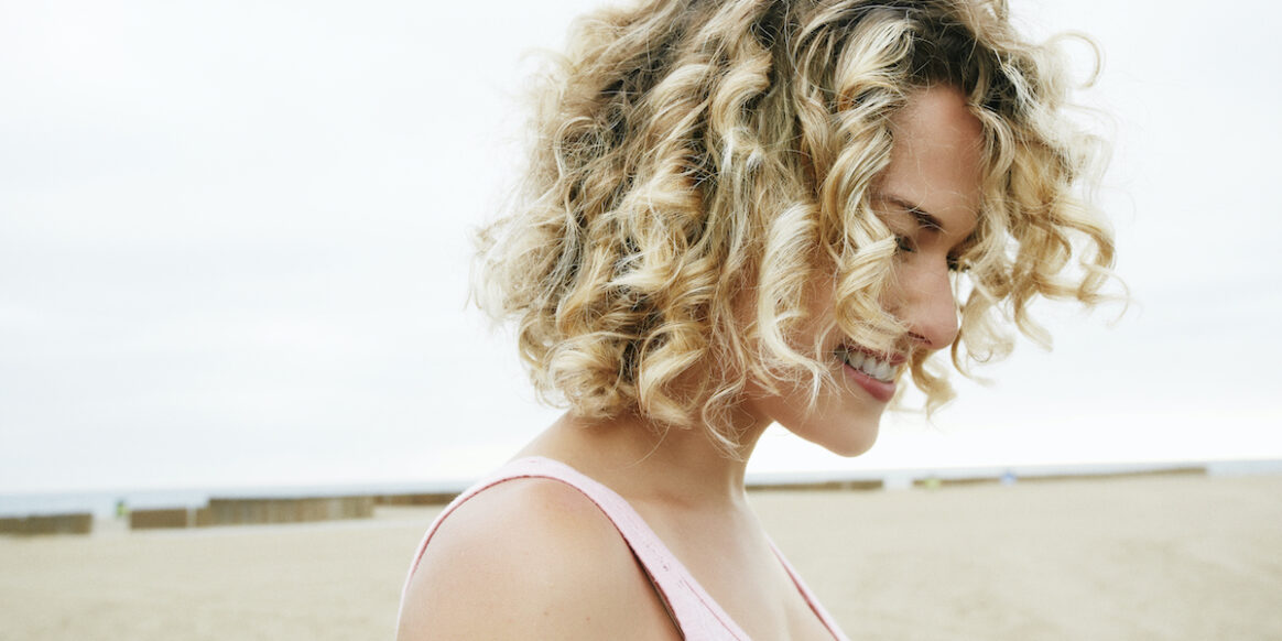 Portrait of smiling young woman with blond curly hair standing on sandy beach.