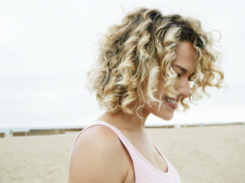 Portrait of smiling young woman with blond curly hair standing on sandy beach.