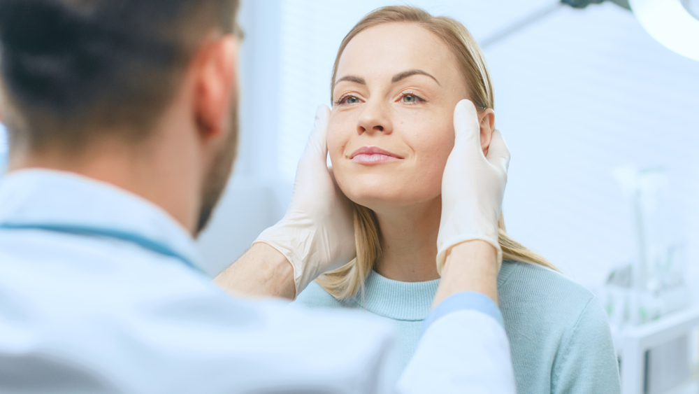 Doctor examining a woman’s face before Sculptra treatment, assessing skin and contours for facial rejuvenation and collagen restoration.