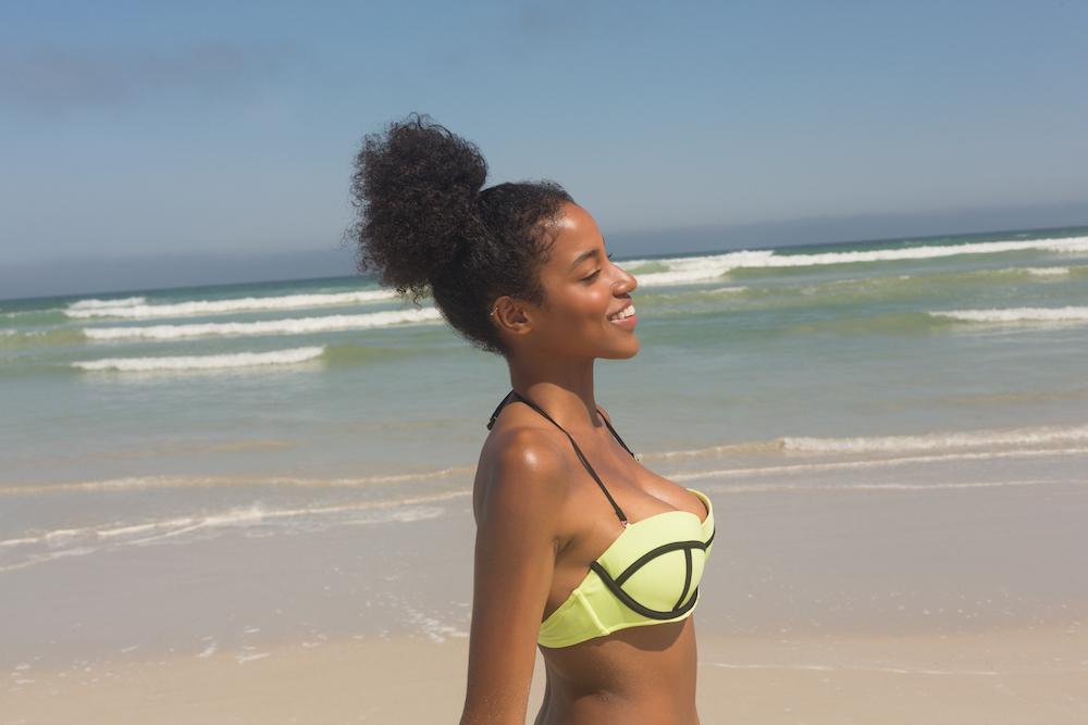 Side view of pretty happy young African American woman in green bikini standing on the beach. She is smiling