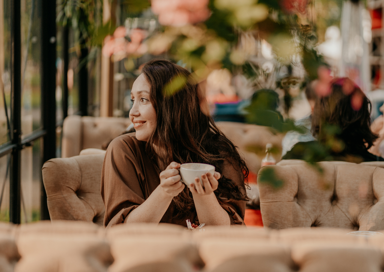 Charming brunette woman with long curly hair sitting at window in cafe with cup of coffee in hands