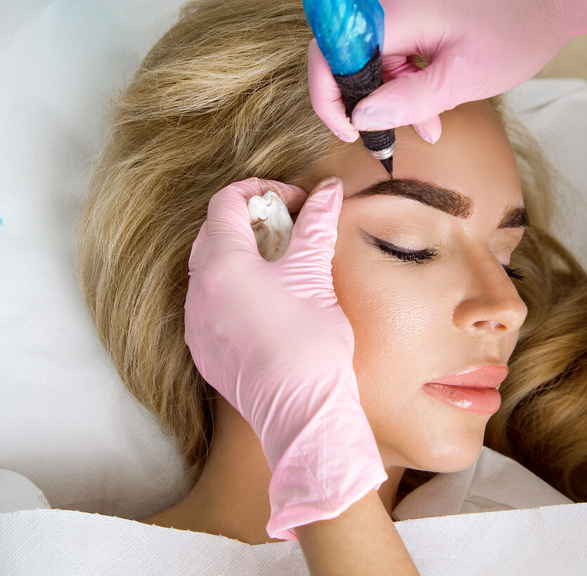 Technician performing a precise brow tint and shaping treatment on a woman lying comfortably with eyes closed.