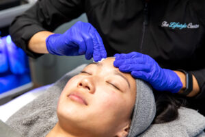 Professional aesthetician performing a facial treatment on a relaxed client, gently massaging the forehead while wearing blue gloves at Aestheticare STL.